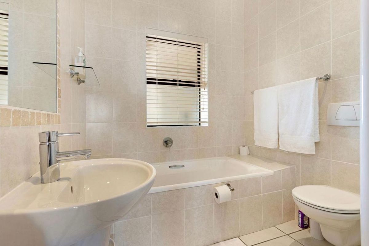 A clean, neutral-toned bathroom featuring a built-in bathtub, pedestal basin, wall-mounted toilet, and full-height wall tiling.
