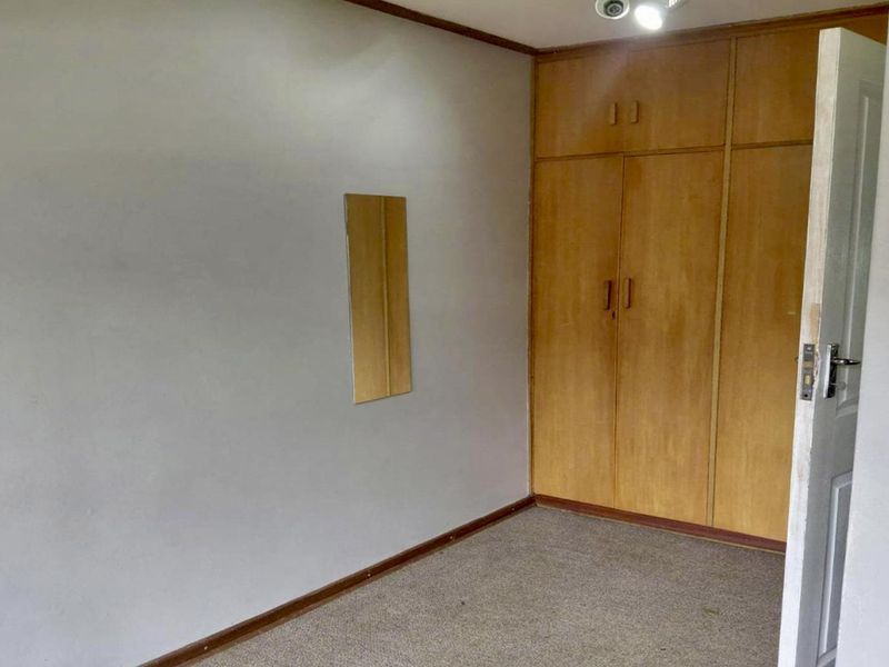 An empty bedroom featuring built-in wooden cupboards, carpeted flooring, and a wall-mounted mirror, presenting a blank canvas for potential updates.