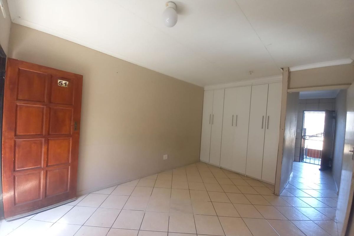 An empty room featuring built-in white cabinetry, tiled flooring, and a wooden entrance door, leading to a hallway with access to a balcony.