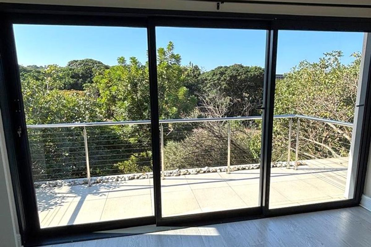 Bright bedroom featuring large sliding glass doors that open onto a private balcony with a stainless steel balustrade and scenic treetop views.
