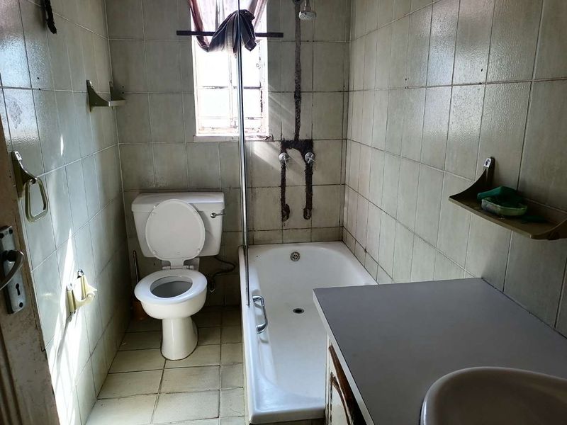 A dated bathroom featuring a standard toilet, bathtub with shower mixer, and a vanity basin, requiring significant renovation and modernization of finishes.
