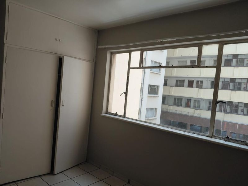 An empty bedroom featuring built-in cupboards, tiled flooring, and large metal-framed windows overlooking the surrounding urban apartment complex.