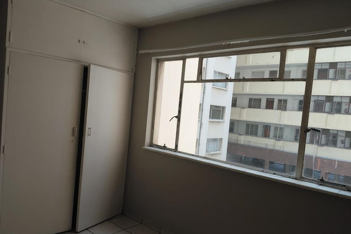 An empty bedroom featuring built-in cupboards, tiled flooring, and large metal-framed windows overlooking the surrounding urban apartment complex.