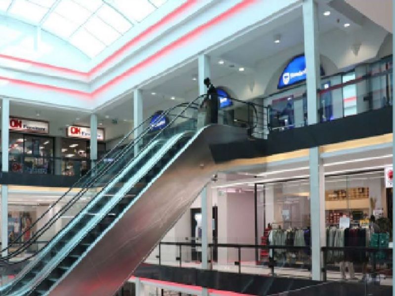 Interior view of a multi-level retail shopping center featuring a central escalator, glass balustrades, and a large overhead skylight.