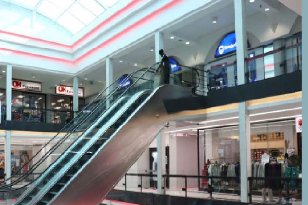 Interior view of a multi-level retail shopping center featuring a central escalator, glass balustrades, and a large overhead skylight.
