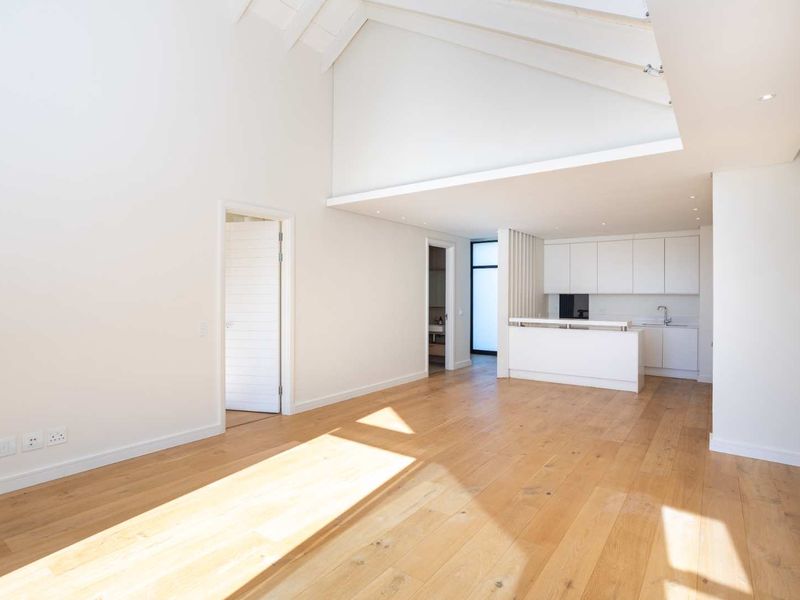 Spacious open-plan living area featuring high vaulted ceilings with exposed rafters, light hardwood flooring, and a modern, minimalist kitchen with built-in cabinetry.