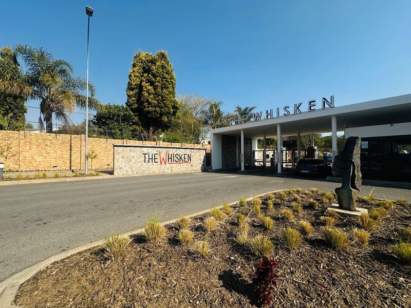 The secure entrance and gatehouse of The Whisken apartment complex, featuring modern signage, landscaped gardens, and perimeter security fencing.