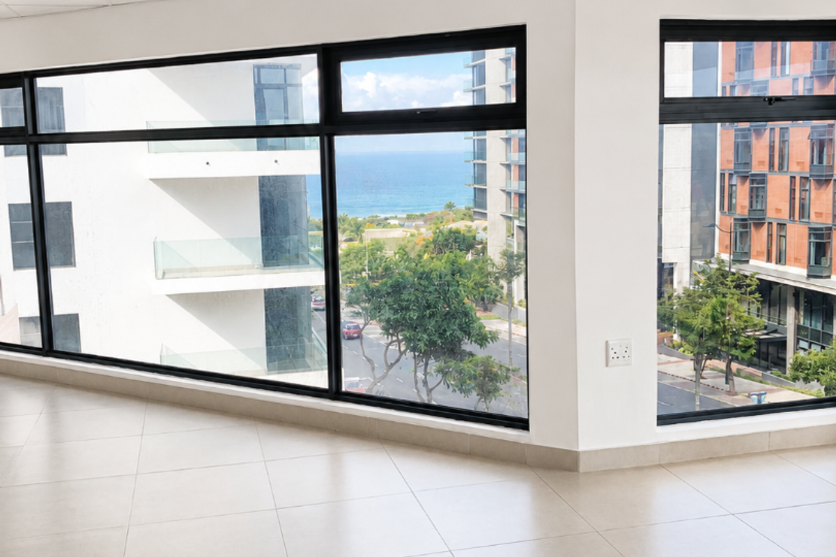 A bright, modern, empty office space featuring large windows with scenic coastal views, neutral tiled flooring, and a clean, professional suspended ceiling.