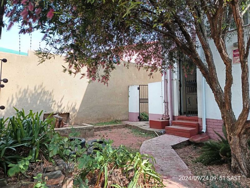 A rear exterior view of the townhouse featuring a small garden area, paved pathway, concrete steps, and perimeter boundary wall with security fencing.