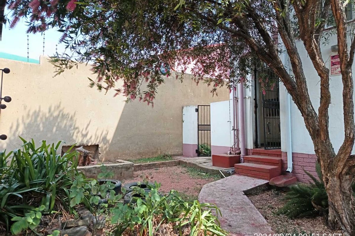 A rear exterior view of the townhouse featuring a small garden area, paved pathway, concrete steps, and perimeter boundary wall with security fencing.