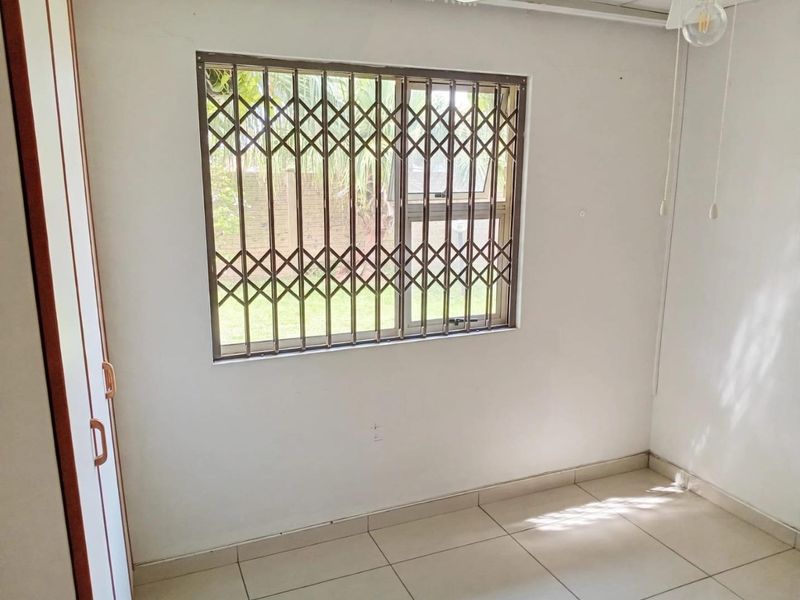 A bright, empty bedroom featuring tiled flooring, a built-in wardrobe, and a window fitted with a security gate.