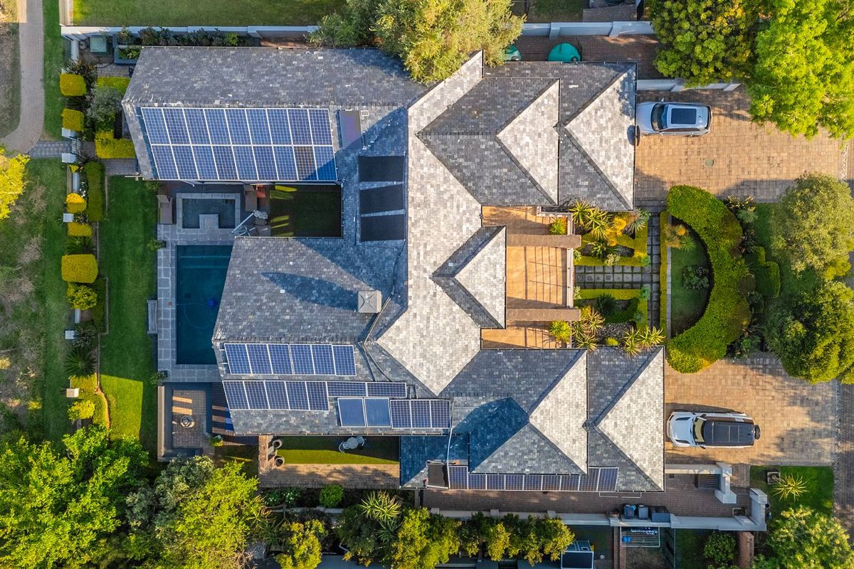 Aerial view of a residential property featuring a large tiled roof with extensive solar panel installation, a swimming pool, and landscaped garden areas.