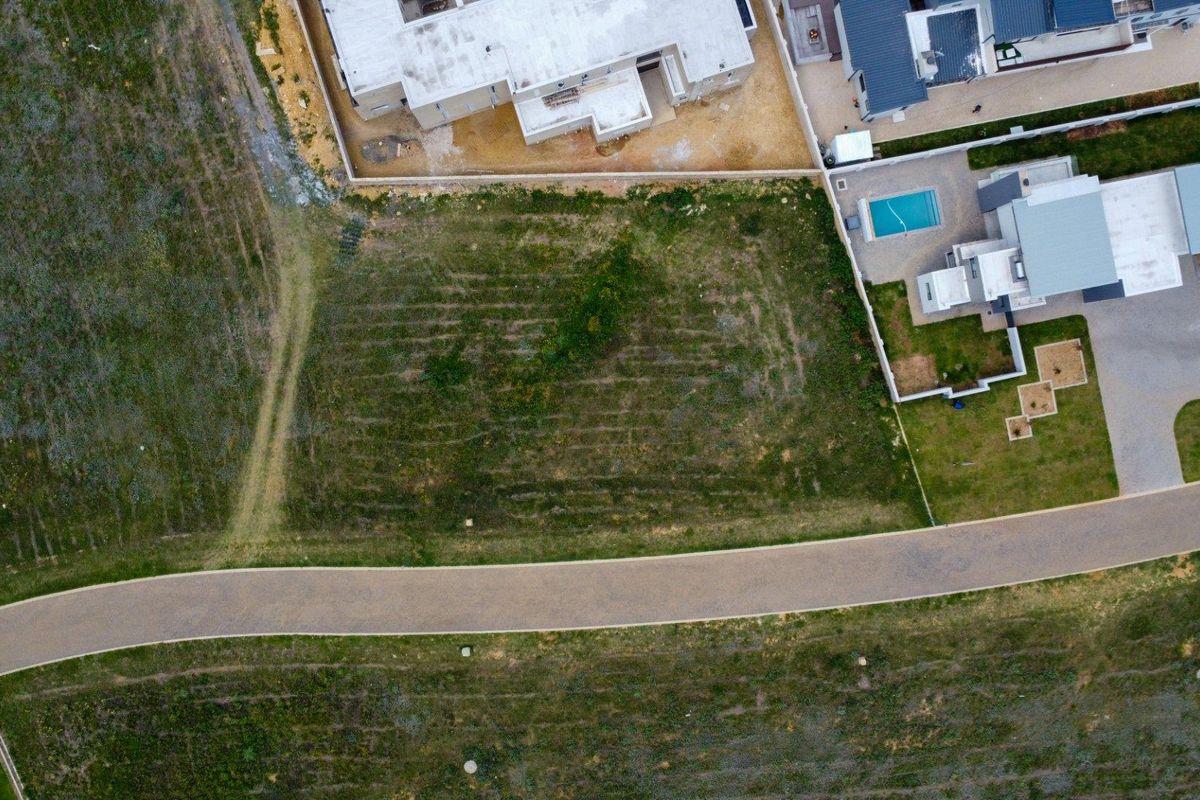 An aerial view of a vacant residential plot situated next to an established paved road and neighboring properties.