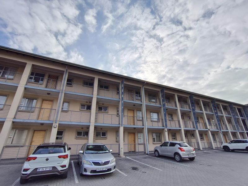 Exterior view of a multi-story apartment complex featuring private balconies, security gates, and a paved parking area for residents.