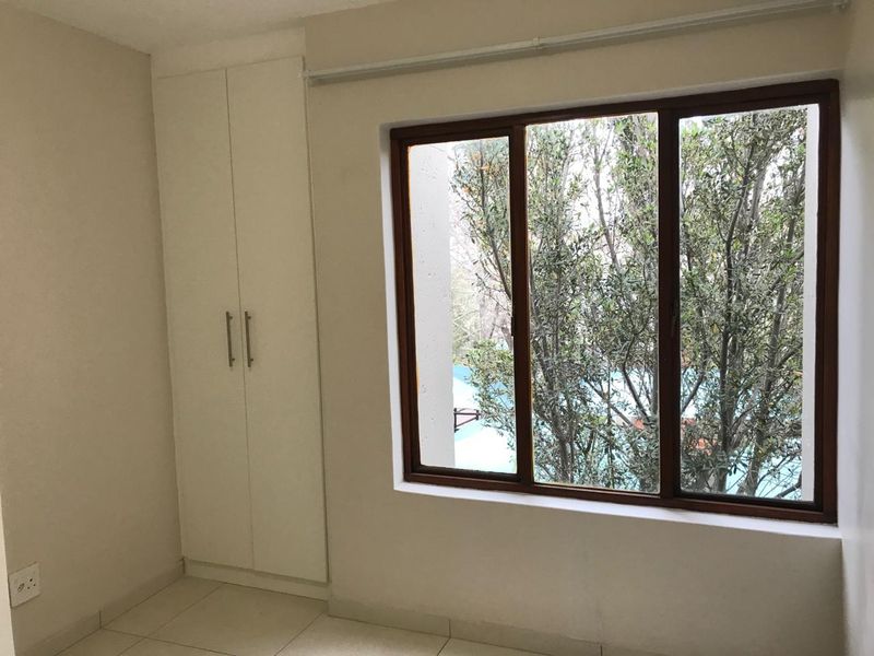 A bright, empty bedroom featuring tiled flooring, a built-in wardrobe, and a large timber-framed window overlooking greenery.