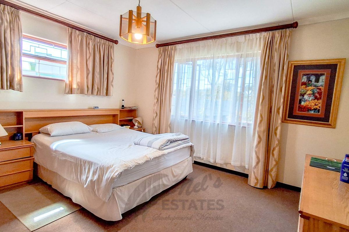A carpeted bedroom featuring a built-in headboard with integrated bedside tables, large windows with security bars, and neutral wall finishes.
