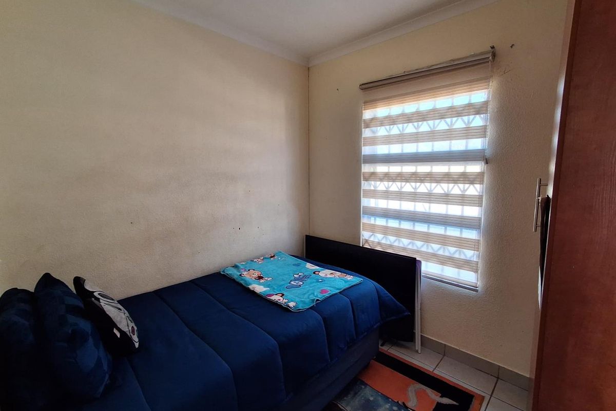 A functional bedroom featuring tiled flooring, a built-in wardrobe, and a window fitted with roller blinds and security bars.