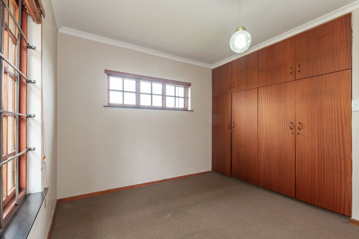 A vacant bedroom featuring wall-to-wall built-in wooden cupboards, carpeted flooring, and a window fitted with security bars.