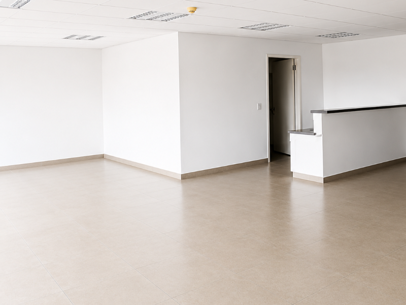 A bright, open-plan office space featuring neutral tiled flooring, a suspended ceiling with recessed lighting, and a built-in reception counter.