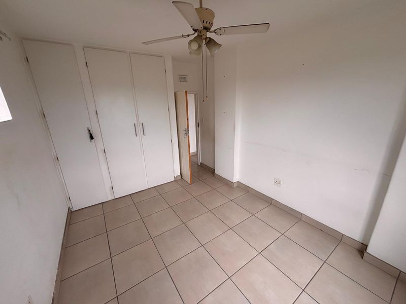 An empty bedroom featuring neutral tiled flooring, built-in cupboards, and a ceiling fan, offering a blank canvas for personalization.