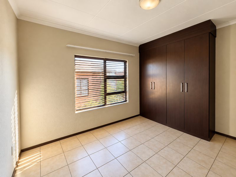 A bright, empty bedroom featuring neutral tiled flooring, built-in dark wood cupboards, and venetian blinds on the window.