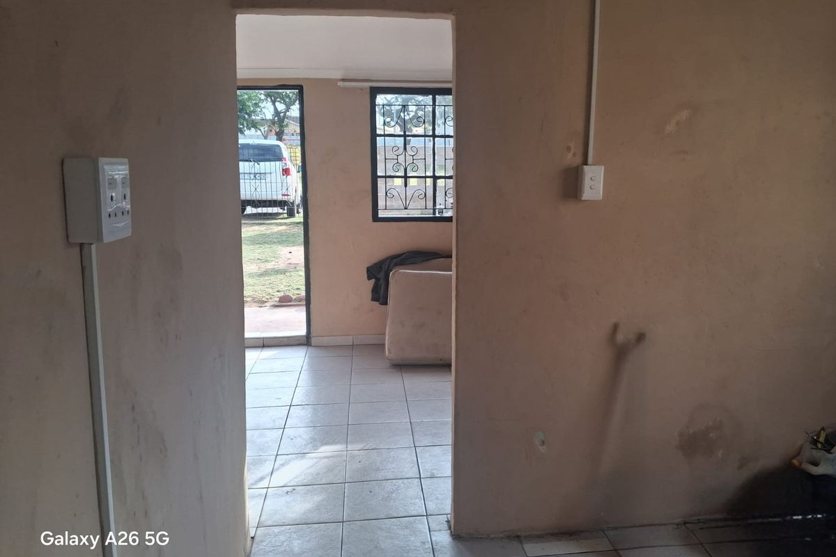 An empty living area featuring tiled flooring, security bars on windows, and visible surface-mounted electrical conduits, requiring cosmetic updates and wall repairs.