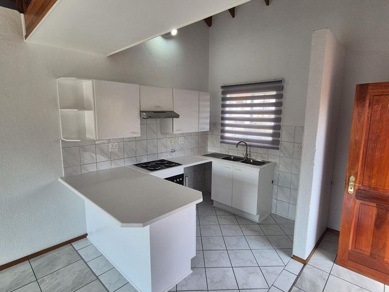 A clean, functional kitchen featuring white cabinetry, a built-in hob with extractor, tiled backsplash, and neutral floor tiles, situated beneath a wooden staircase.