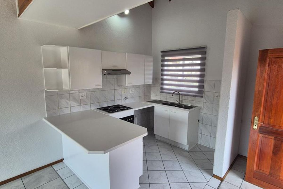 A clean, functional kitchen featuring white cabinetry, a built-in hob with extractor, tiled backsplash, and neutral floor tiles, situated beneath a wooden staircase.