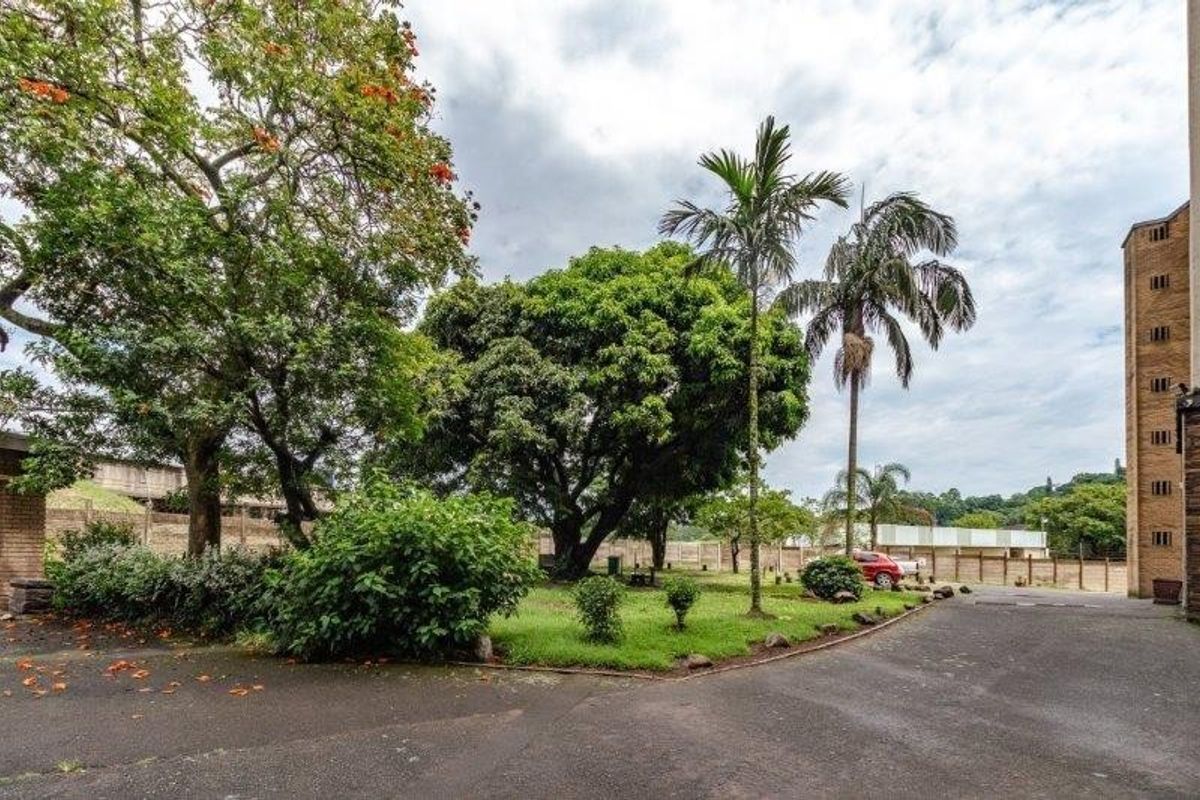 A view of the apartment complex exterior featuring a paved driveway, established garden area with mature trees, and perimeter boundary walling.