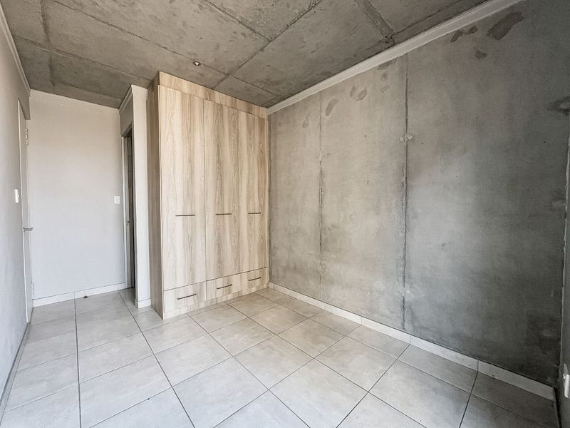 A minimalist bedroom featuring exposed concrete walls and ceiling, tiled flooring, and a modern built-in wooden wardrobe.