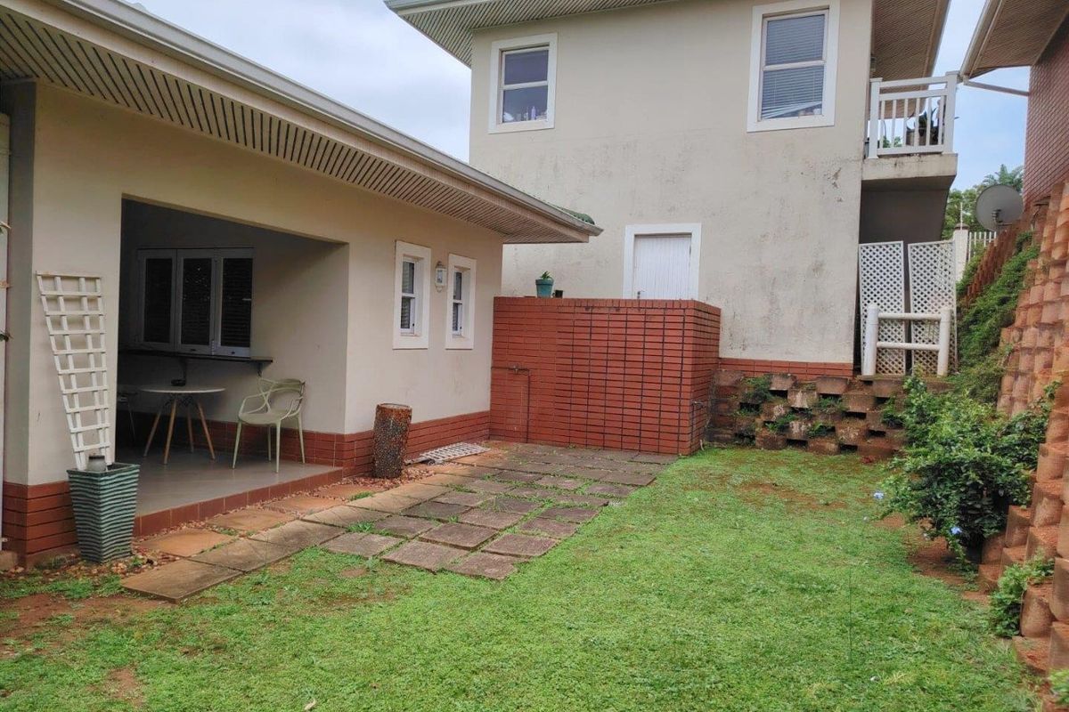 Rear exterior view featuring a covered patio, paved walkway, lawn area, and a brick retaining wall leading to the upper level of the house.