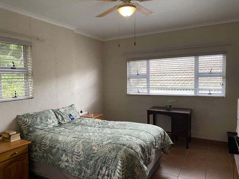 A bedroom featuring tiled flooring, a ceiling fan, and dual windows with venetian blinds. The walls show signs of wear and require maintenance.