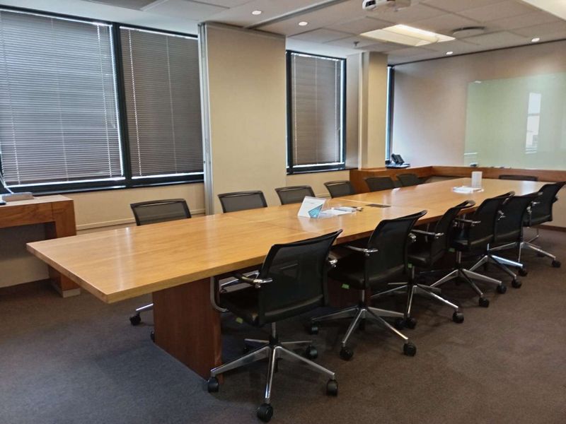 A professional boardroom featuring a large wooden conference table, suspended ceiling with recessed lighting, and large windows with venetian blinds.