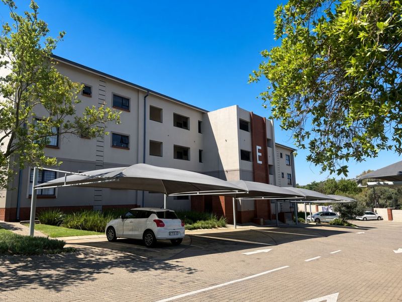 Exterior view of a modern multi-story apartment building featuring paved parking areas with shade cloth carports and well-maintained landscaped garden beds.