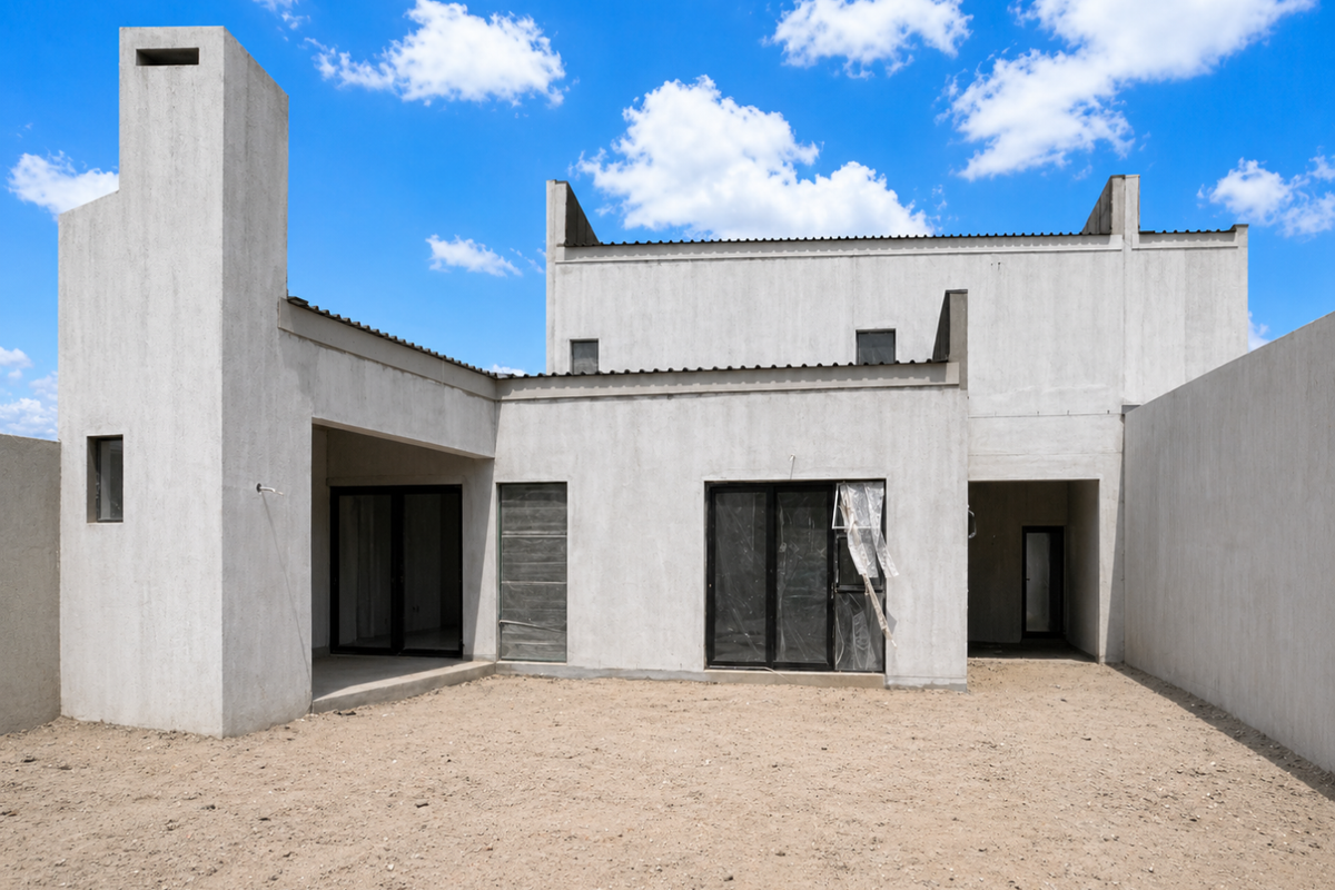 Rear view of a modern architectural house under construction, featuring a prominent chimney, sliding glass doors, and an expansive unfinished backyard area.
