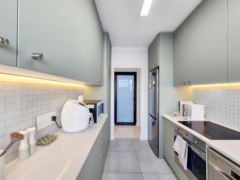 Modern galley kitchen featuring sage green cabinetry, stone countertops, mosaic tile backsplash, under-cabinet lighting, and high-quality integrated appliances.