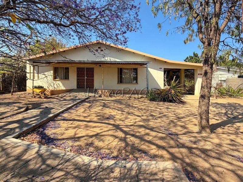 Front view of a single-story house featuring a covered patio, security gate, and an attached carport, set on a large, unlandscaped plot.