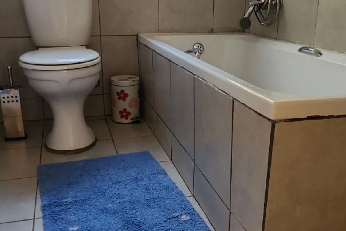 A functional bathroom featuring a tiled bathtub, toilet, and large window with security bars, finished with neutral ceramic wall and floor tiles.