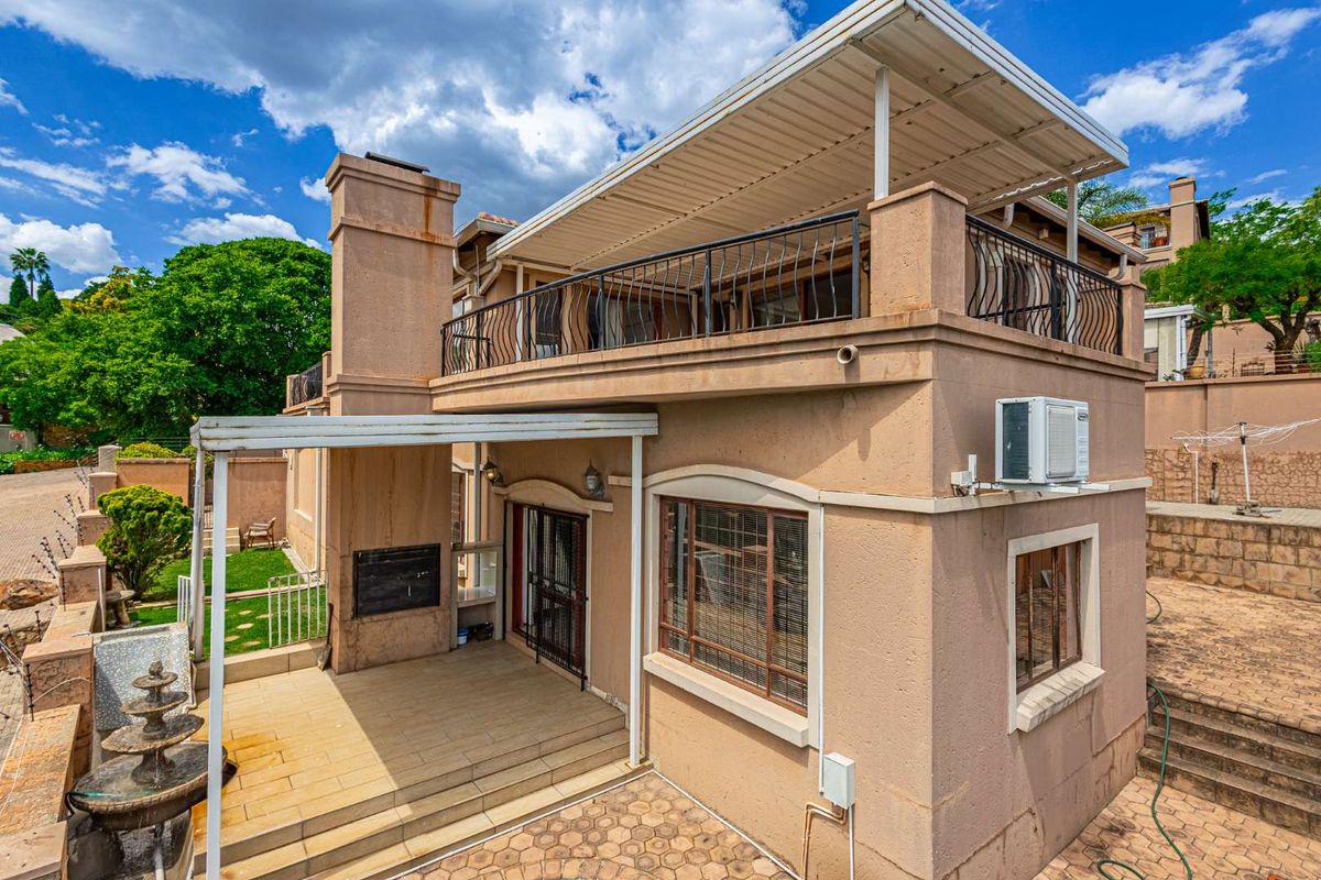 Rear exterior view featuring a covered patio with built-in braai, an upper-level balcony with wrought iron railings, and paved outdoor areas.