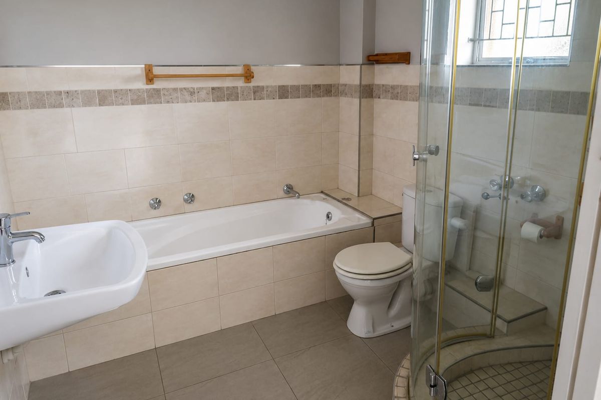 A functional bathroom featuring a built-in bathtub, a separate glass-enclosed shower, a pedestal basin, and a toilet with neutral wall and floor tiling.