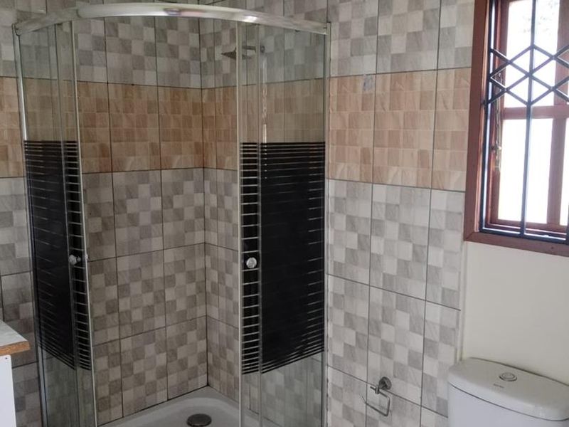 A functional bathroom featuring a corner shower enclosure, a standard toilet, and decorative wall tiling with a window fitted with security bars.