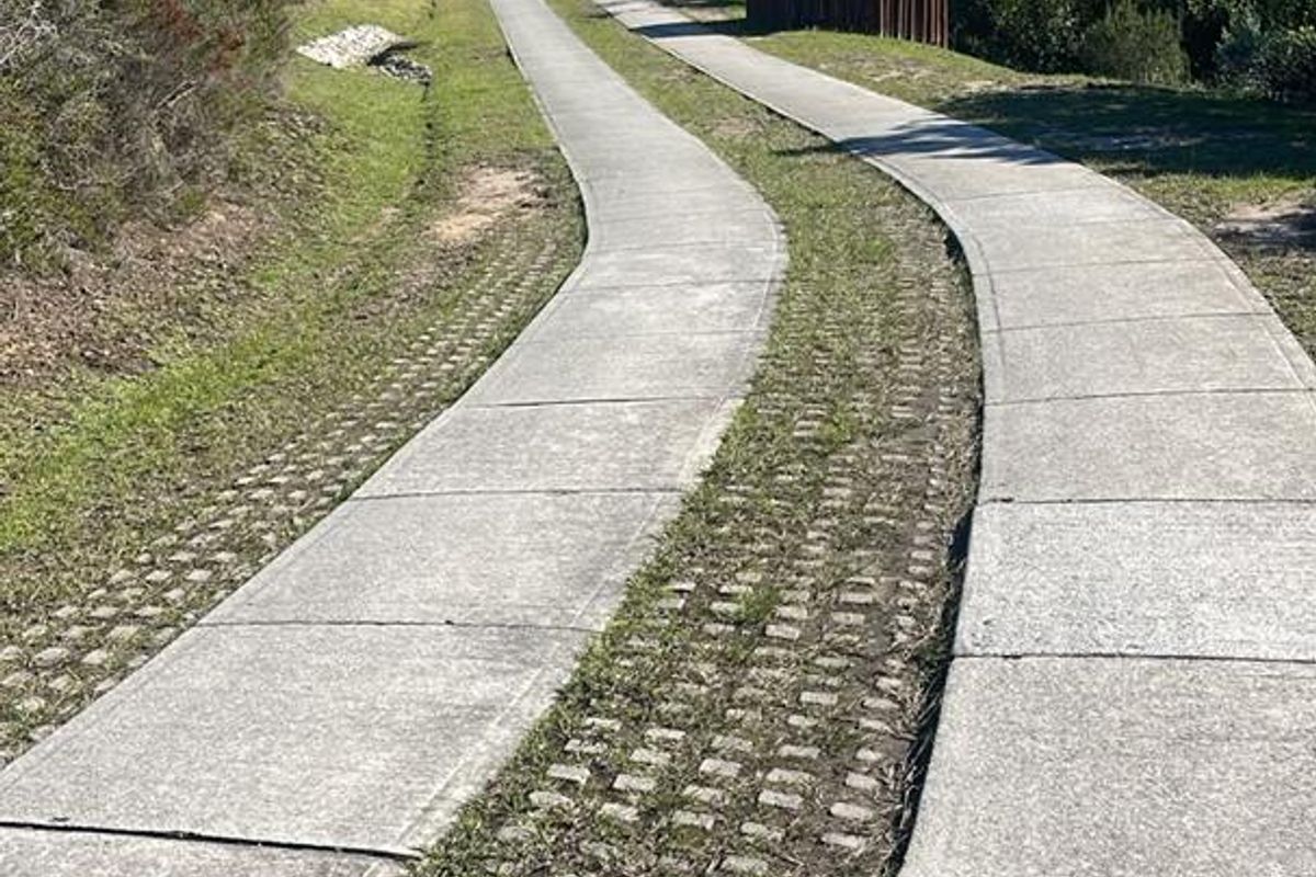 A paved concrete driveway with grass paver center strip leading through a landscaped residential plot with natural vegetation.