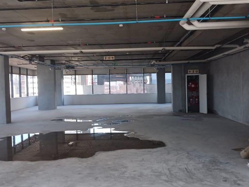 An open-plan office space in shell condition featuring exposed concrete ceilings, large windows, and visible utility piping, currently requiring significant interior fit-out.