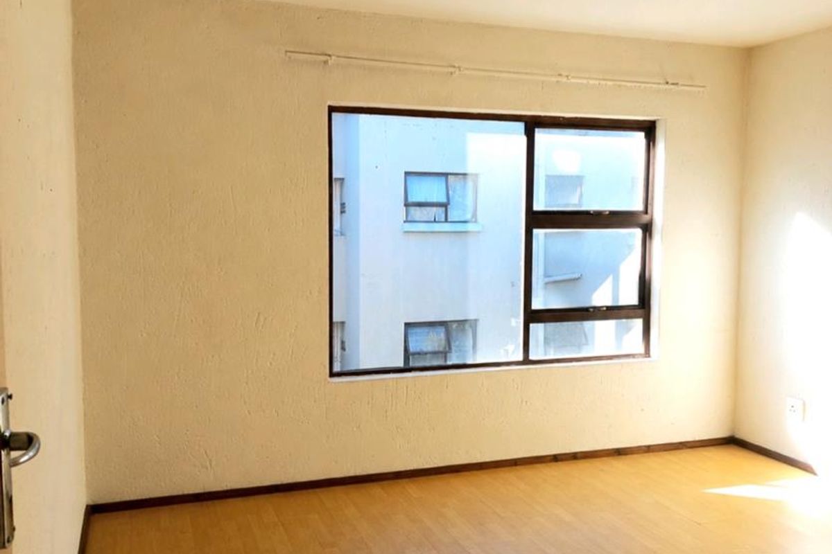 A bright, empty bedroom featuring laminate flooring, neutral walls, and a large window with an aluminium frame.