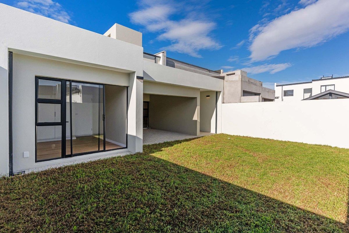 A modern rear exterior view featuring a covered patio, sliding glass doors, and a private lawn area enclosed by boundary walls.