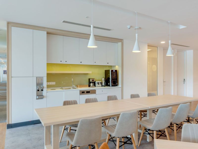 Modern office kitchenette featuring sleek white cabinetry, integrated appliances, a glass splashback, and contemporary pendant lighting, adjacent to a communal dining area.