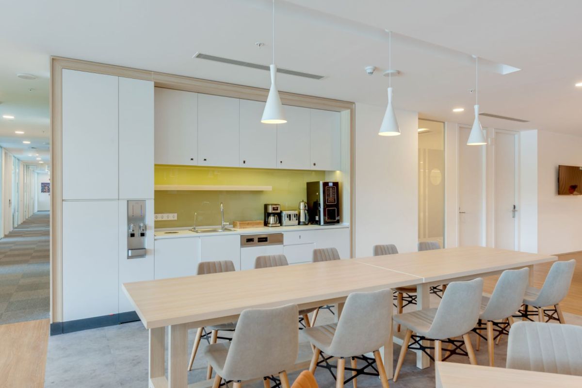 Modern office kitchenette featuring sleek white cabinetry, integrated appliances, a glass splashback, and contemporary pendant lighting, adjacent to a communal dining area.