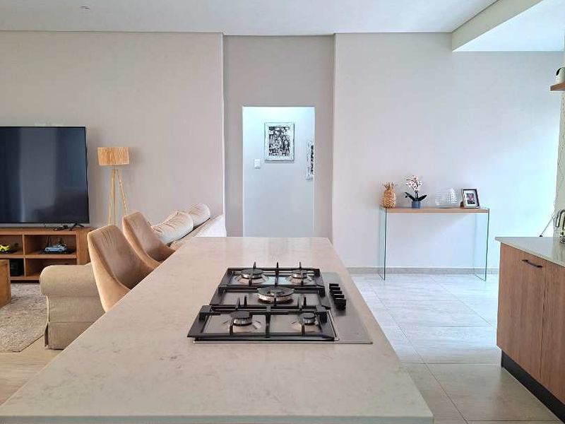 Modern open-plan living area featuring a central kitchen island with a built-in gas hob, recessed ceiling lighting, and neutral tiled flooring throughout.