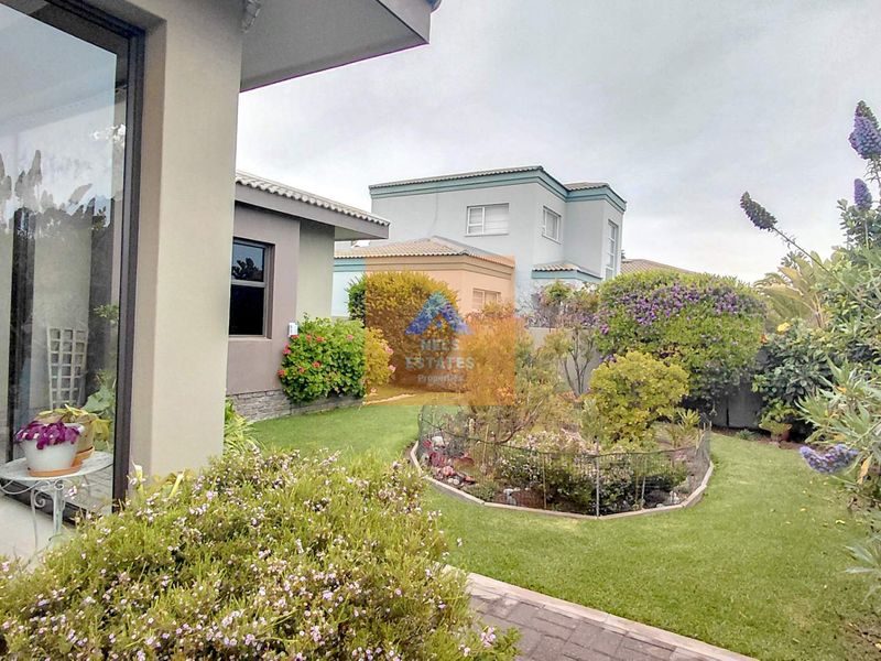 A well-maintained rear garden featuring a landscaped flower bed, lush lawn, and paved walkway, accessible via large sliding glass doors.