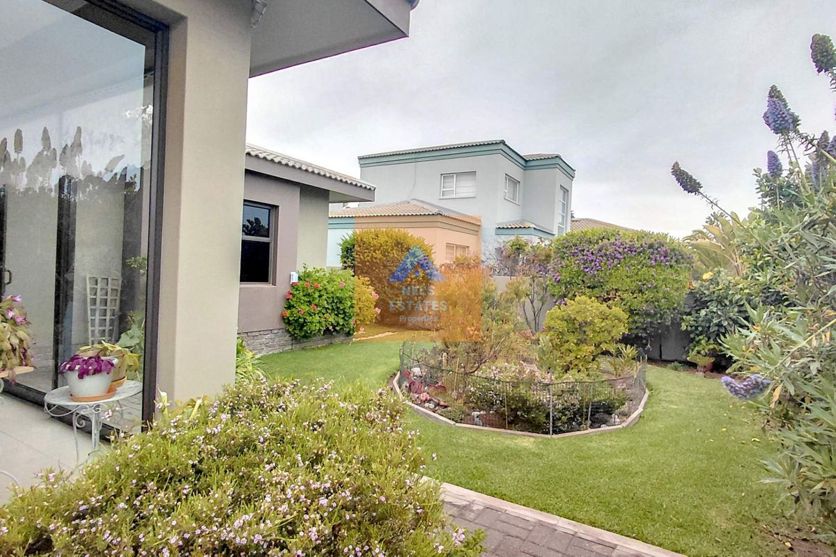 A well-maintained rear garden featuring a landscaped flower bed, lush lawn, and paved walkway, accessible via large sliding glass doors.